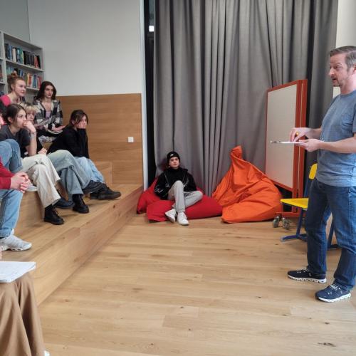 Schuler:innengruppe sitzt in einer Bibliothek auf Stufen, rechts erklärt Markus Koschuh etwas