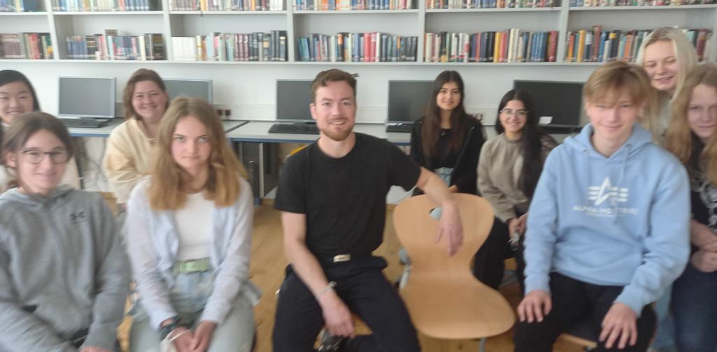 Ein Mann und Schüler:innen sitzen vor einer Bücherwand