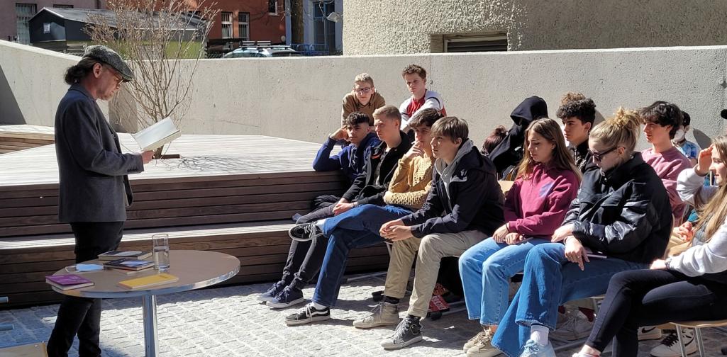 Ein Mann liest stehend aus einem Buch, rechts sitzen Schüler:innen und hören zu