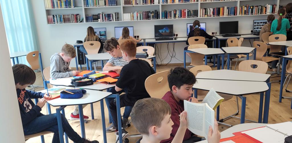 Einige Schüler:innen beim Lesen an Tischen und an Computern im Hintergrund